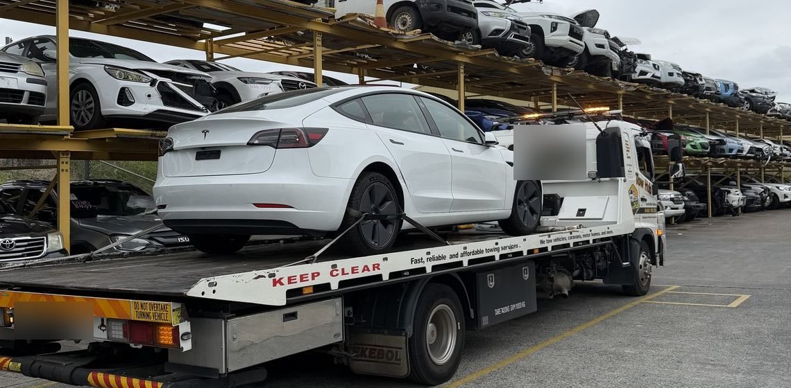 Flatbed tow truck loaded with a white Tesla at a salvage yard